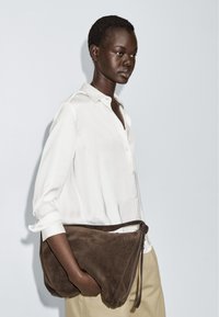Brown suede shoulder bag with a curved shape, zipper closure, and long leather strap, held underarm against a white background.