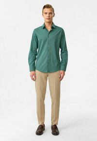 Groene overhemd met lange mouwen en knopen, gemaakt van een gladde stof, gecombineerd met beige broeken en bruine loafers, tentoongesteld op een neutrale achtergrond.