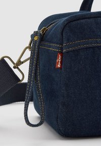 Dark blue denim bag with a rectangular shape, featuring gold stitching, a webbed strap, and a Levi's logo tag. Hardware is brass-colored.