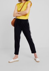 Svarta joggerbyxor med vita sidostråkar, åtföljda av en gul kortärmad topp; modellen bär vita espadriller och har en brun axelväska.