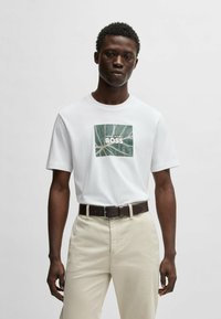 Camiseta blanca de algodón con un diseño de hoja verde en forma de cuadrado que lleva el texto "BOSS". Combinada con pantalones de color claro y un cinturón marrón oscuro.