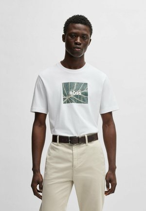 T-shirt en coton blanc avec un design de feuille verte carrée et le texte "BOSS". Associé à un pantalon de couleur claire et une ceinture marron foncé.