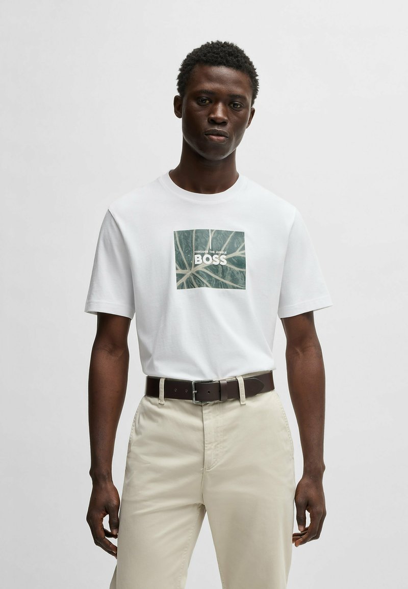 Camiseta blanca de algodón con un diseño de hoja verde en forma de cuadrado que lleva el texto "BOSS". Combinada con pantalones de color claro y un cinturón marrón oscuro.