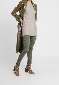 Trench-coat brillant vert olive sur un haut en tricot beige, associé à un pantalon cargo olive et des bottines à talon carré en imprimé serpent marron.