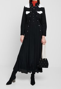 Abito nero con un design fluido e maniche lunghe, indossato sotto una camicia nera e bianca con bottoni. Stivali neri con tacco e una borsa nera.