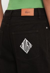 Κοντινό πλάνο πίσω τσέπης μαύρου παντελονιού με λευκή κεντητή γραφή "LACOSTE" σε σχήμα διαμαντιού και καφέ δερμάτινη ετικέτα με λογότυπο κροκόδειλου επάνω.