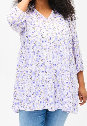 Kvinde iført en løs, langærmet hvid tunika med et lilla blomsterprint og lyseblå jeans, stående mod en ensfarvet baggrund.