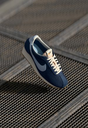 Laivastonsininen Nike-tennari, jossa valkoinen swoosh-merkki ja nauhat, lepää teksturoidulla metallipinnalla luonnonvalossa.