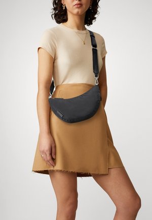Borsa a tracolla nera in nylon con design curvo, tracolla regolabile e hardware discreto. Indossata con un top beige a coste e una gonna marrone chiaro.