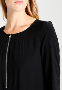 Top nero con scollatura rotonda e chiusura con zip. Presenta dettagli arricciati sulle spalle e un tessuto morbido e liscio.