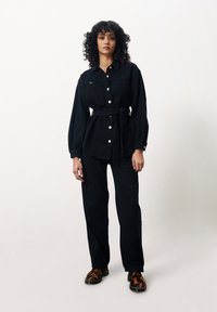 Chemise en denim noire avec fermeture à boutons, taille cintrée et manches longues. Associée à un pantalon noir et des chaussures texturées marron et noires.