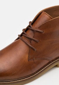 Bottine marron en cuir à lacets avec coutures apparentes et semelle en caoutchouc sur fond blanc.