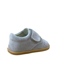 Zapato de bebé de ante gris claro con tira de Velcro y suela de goma marrón, mostrado desde un ángulo lateral trasero sobre un fondo blanco.