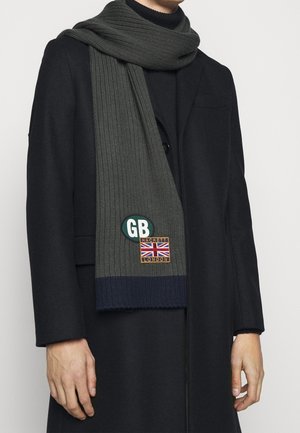 Homme portant un manteau noir et une écharpe tricotée vert foncé avec un patch GB et l'emblème Union Jack Hackett London.