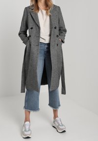 Žena v černobílé kostkované bundě, béžovém svetru, zkrácených modrých džínách s roztrhanými lemy a bílých teniskách, stojící s rukama v kapsách.