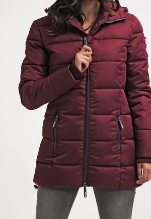 Frau zieht eine lange, gepolsterte, dunkelrote Winterjacke mit Reißverschlusstaschen an den Seiten und Kapuze hoch, trägt graue Hose.