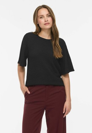 Femme aux longs cheveux bruns portant un haut noir texturé à manches courtes et un pantalon bordeaux, debout avec une main dans la poche.
