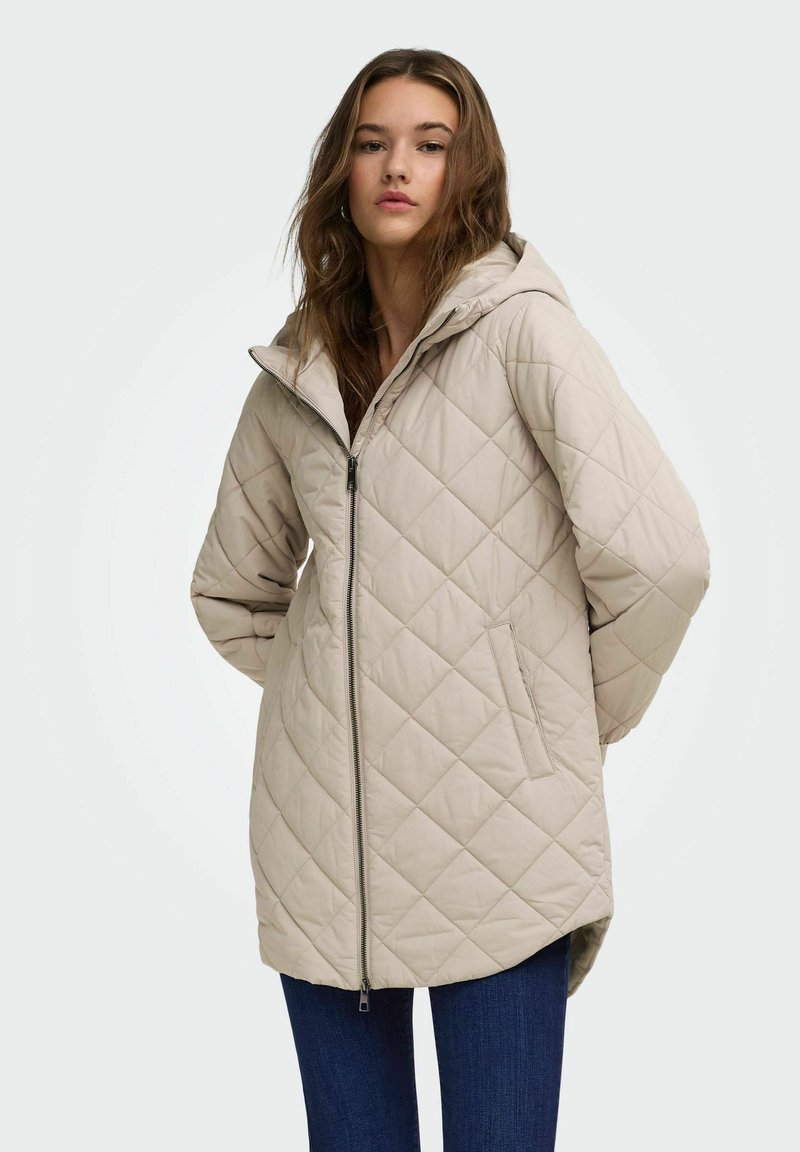 Jeune femme portant une veste matelassée beige à capuche avec fermeture éclair devant et poches, associée à un jean bleu foncé, debout devant un fond clair.