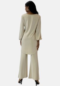 Beige langærmet top med buet kant og bukser med vide ben, lavet af et glat stof. Kombineret med sorte højhælede sko.