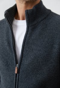 Sweat à zip gris foncé avec un tissu texturé. Présente un col haut et un accent en cuir sur la tirette de la fermeture éclair. Porté par-dessus une chemise blanche.