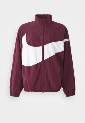 Burgundy színű zipzáros dzseki magas gallérral, nagy fehér Nike swoosh logóval, elasztikus mandzsettákkal és aljjal. Könnyű, texturált anyag.