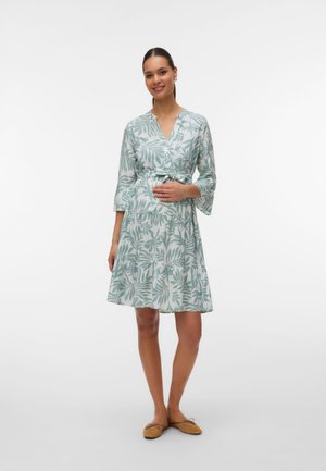 Femme en robe mi-longue verte et blanche à imprimé feuilles, manches trois-quarts, main posée sur le ventre, portant des ballerines beiges.