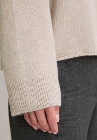 Beżowy sweter dzianinowy z prążkowanymi mankietami, w połączeniu z ciemnoszarymi spodniami w kratkę. Sweter ma miękką teksturę i swobodny krój.