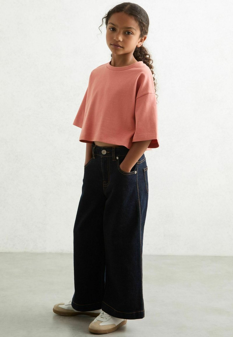 Rózsaszín pamut rövid ujjú crop top, széles szárú sötét farmernadrággal párosítva. Szabványos zsebekkel és fehér cipőkkel kiegészítve.