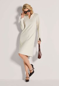 Frau in einem cremefarbenen, langärmeligen Kleid bis zum Knie, mit schwarzen Absätzen und Sonnenbrille, die eine braune handgefertigte Tasche vor einem einfarbigen Hintergrund hält.