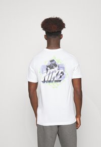 Camiseta de algodón blanca con un gráfico colorido en la parte posterior, que presenta "NIKE" en texto negrita, contorneado en verde sobre un diseño dinámico.