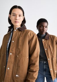 Brune jakker med fløjlskraver, trykknaplukninger og forlommer; båret over grå og denim beklædning. To modeller vender fremad.