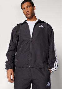 Μαύρο αθλητικό μπουφάν με φερμουάρ μπροστά, ριγωτούς τριγμούς και άσπρες ρίγες στα μανίκια. Διαθέτει λογότυπο στο στήθος και τσέπες στις πλευρές.