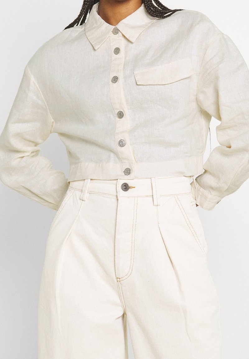 Femme portant une chemise courte écrue à boutons avec manches longues et un pantalon large taille haute de couleur similaire.