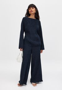 Conjunto de dos piezas texturizado en color azul marino que incluye una blusa de manga larga con cuello barco y pantalones de pierna ancha, acompañado de un bolso tejido blanco.