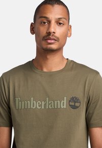 Camiseta de algodón verde olivo con cuello redondo, que presenta un gran logotipo en verde claro de "Timberland" y un gráfico de árbol en la parte frontal.