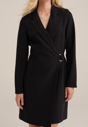 Kobieta w czarnej sukience marynarkowej do kolan z długimi rękawami i ozdobnym złotym pierścieniem zapinającym w talii.
