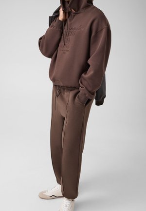 Personne portant un sweat à capuche marron et un pantalon de survêtement assorti avec des baskets blanches, debout avec une main dans la poche.