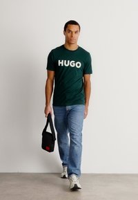 Camiseta de manga curta verde-escuro com grande impressão branca "HUGO", jeans azul claro, bolsa transversal preta e ténis brancos sobre um fundo simples.