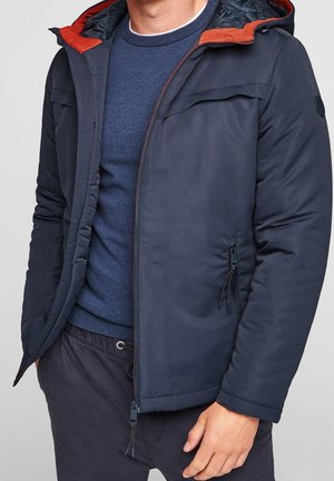 Homme portant une veste matelassée bleu marine avec une doublure orange sur un pull bleu, avec des poches zippées et un pantalon à cordon.