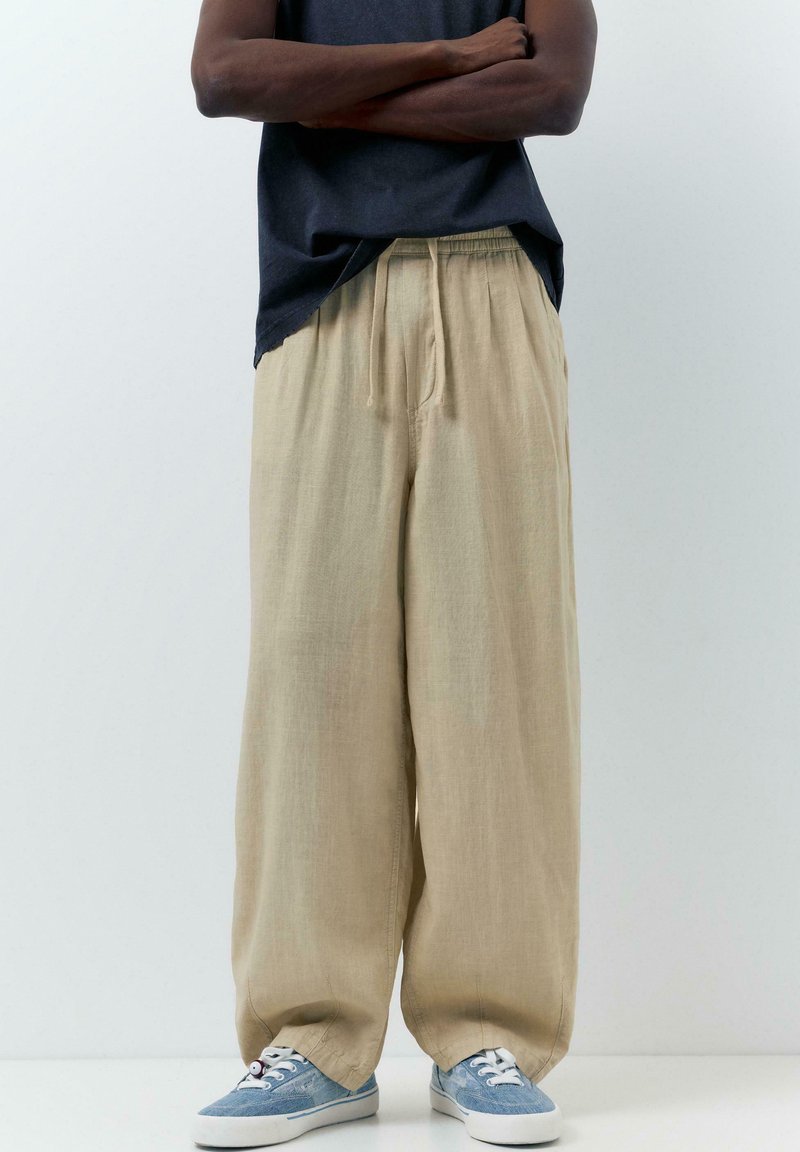 Personne portant un pantalon beige à jambes larges, des baskets en toile bleues et un haut noir, les bras croisés, sur un fond blanc uni.