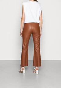 Vrouw in hooggeritste bruine leren broek, witte mouwloze top en witte haksandalen, staand tegen een effen lichte achtergrond.