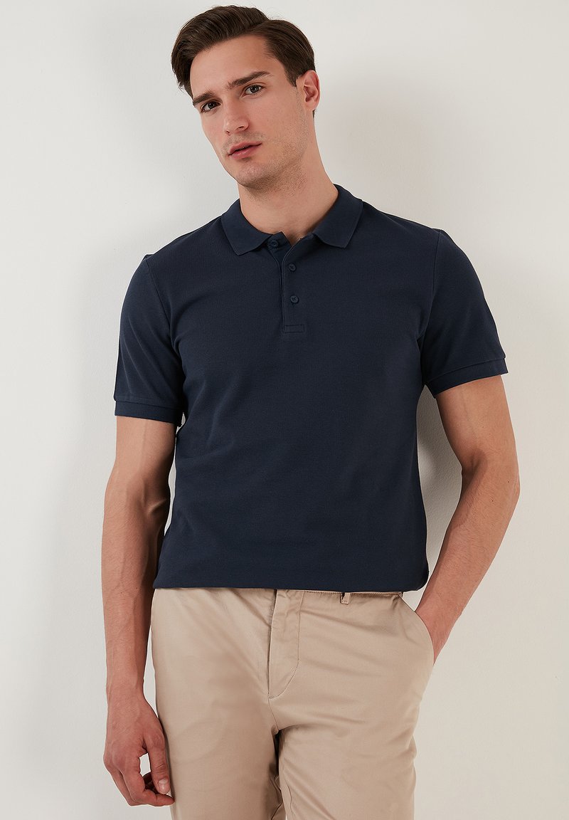 Polo de color azul marino en mezcla de algodón, con cuello clásico, botonera de tres botones y mangas cortas. Llevado con pantalones beige.