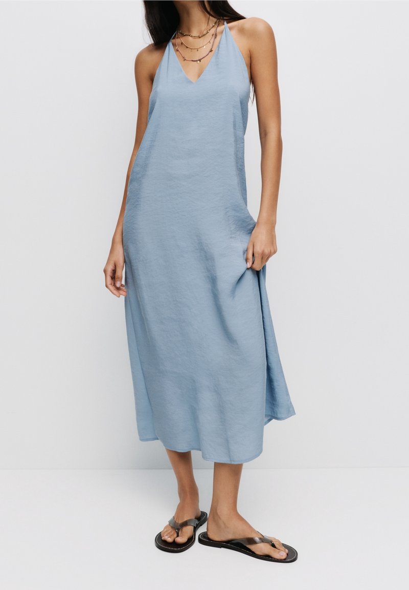 Femme portant une robe midi bleu clair sans manches avec un col en V et des sandales noires, debout devant un fond blanc uni.