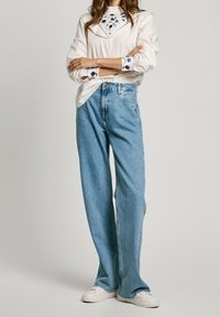 Vit långärmad skjorta med blå mönster, i kombination med ljusblå vida jeans och vita sneakers. Outfitten har detaljerade manschetter.