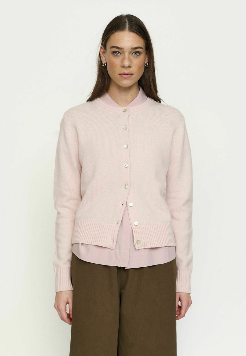Jeune femme aux cheveux bruns et raides, portant un cardigan à boutons rose clair, un chemisier rose et un pantalon marron, debout devant un fond uni.