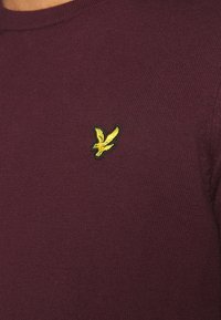 Maglia a maglia di colore bordeaux con scollatura rotonda e un piccolo logo con uccellino ricamato di colore giallo sul lato sinistro del petto. Texture morbida e design aderente.
