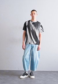 Fekete és fehér absztrakt mintás póló, világoskék, bő szárú farmernadrág, és fekete, fehér talpú sportcipő, egy texturált szürke padlón állva.