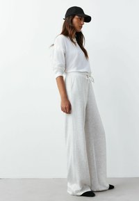 Hellgraue Sweatpants mit weitem Bein und Tunnelzugbund, kombiniert mit einem weißen Langarmshirt und einer schwarzen Mütze, vor einem neutralen Hintergrund positioniert.