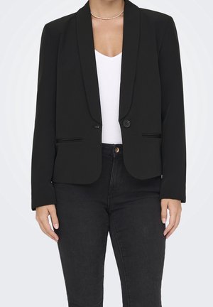 Femme portant un blazer noir ajusté avec un seul bouton, un haut blanc à col en V, un pantalon noir et un collier fin de perles.