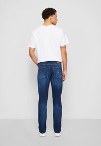 Άνδρας γυρισμένος προς τα πίσω, φορώντας λευκό μπλουζάκι, μπλε τζιν με πίσω τσέπες και λευκά αθλητικά παπούτσια, που στέκεται σε έναν απλό γκρίζο φόντο.